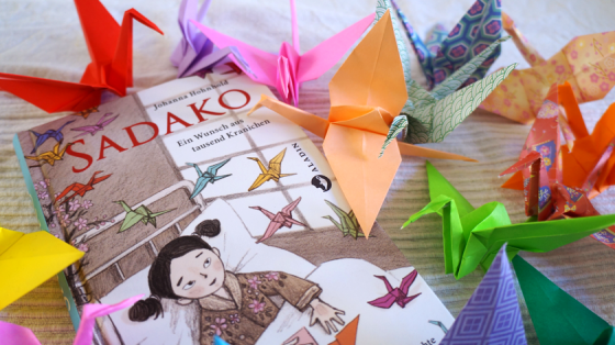 Das Kinderbuch Sadako von Johanna Hohnhold und einige der ikonischen Papierkraniche, die wohl für immer mit Sadakos Schicksal verbunden sein werden.